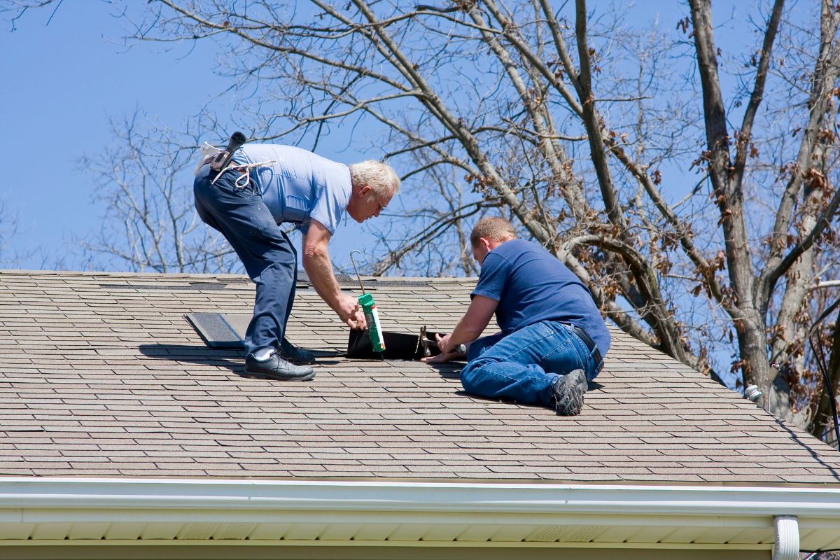 Two workers on a roof are repairing a vent. One holds a caulking gun while the other adjusts the vent in sunny weather.