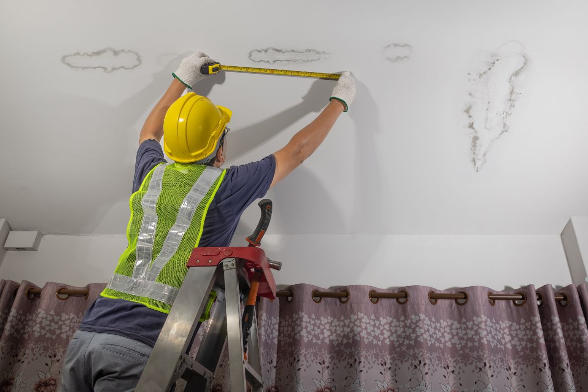 A worker in a yellow hard hat measures ceiling stains on a ladder, wearing a reflective vest and gloves, with curtains in the background.