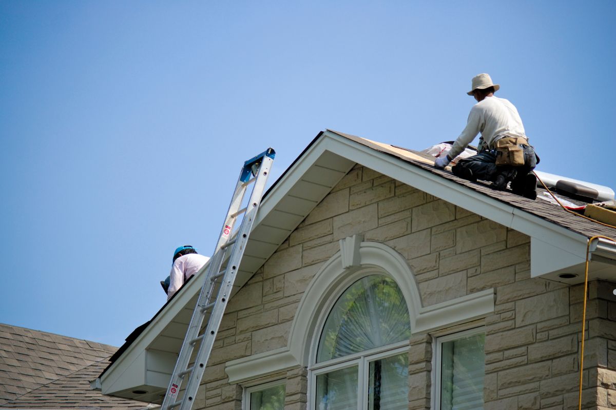 Two roofers working on a sloped roof, one on a ladder and the other installing shingles under a clear blue sky.