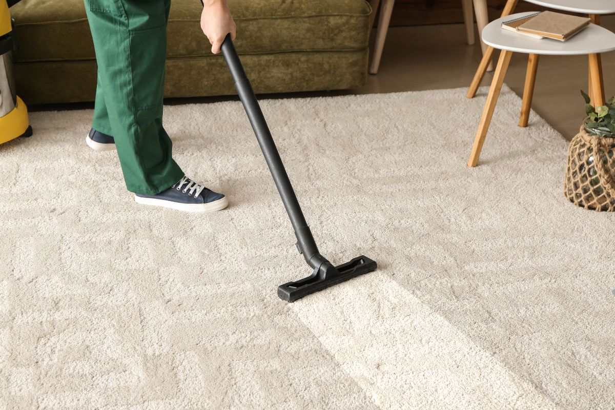 A person in green pants vacuums a light-colored carpet with a black vacuum cleaner in a cozy living room setting.