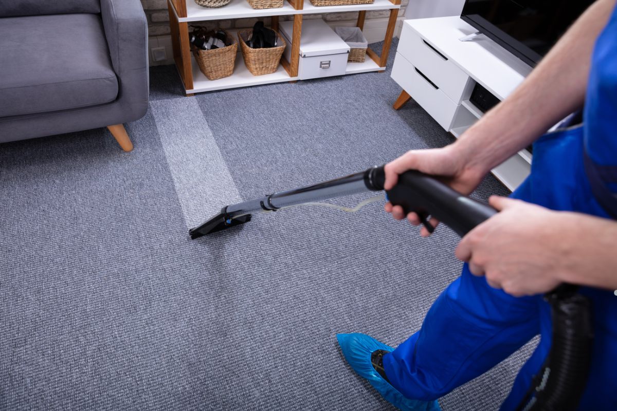 A person in blue attire cleans a gray carpet with a vacuum cleaner, revealing a clean strip against the uncleaned area.